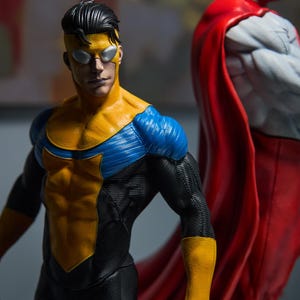 May include: Two action figures of superheroes. The first figure is in a yellow and blue suit with a black undersuit. The second figure is in a white and red suit with a red cape. The figures are highly detailed and posed in a dynamic stance.