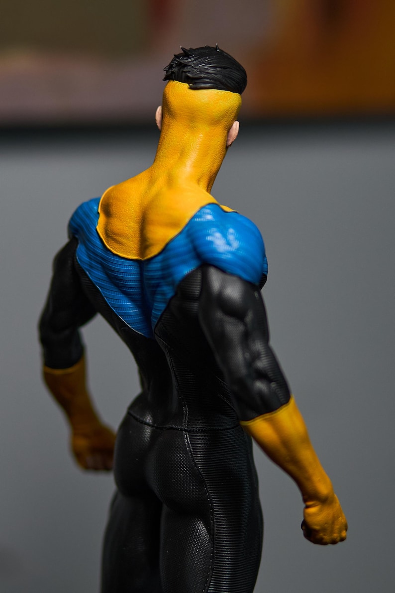 May include: A figurine of a superhero from the back, showcasing a yellow head and shoulders, blue and black suit. The figure has a muscular physique and is posed in a dynamic stance. The colors are vibrant and the details are well-defined.