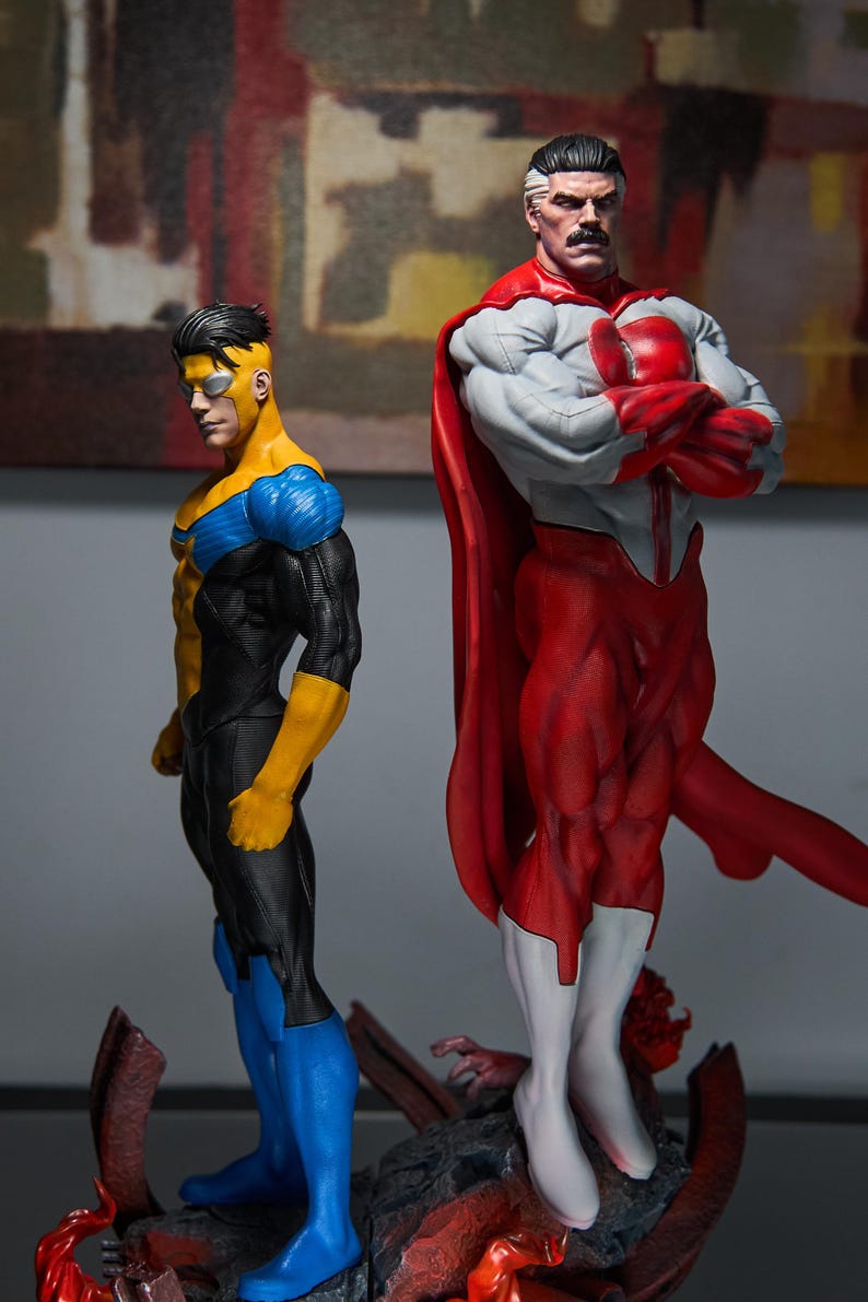 May include: Two superhero action figures. One figure is in a yellow, blue, and black suit. The other figure is in a red and white suit with a red cape. Both figures are standing on a rocky base.