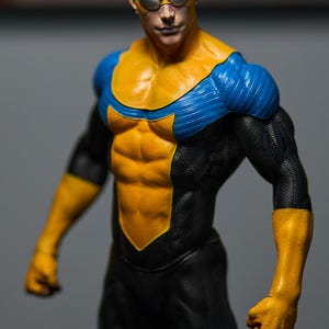 May include: A detailed action figure of a superhero in a dynamic pose. The figure is clad in a black, yellow, and blue costume. The yellow accents highlight the chest, arms, and gloves, while blue accents are on the shoulders. The figure has a determined expression.