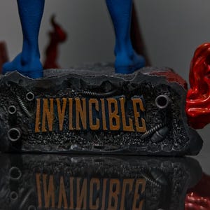 May include: A detailed statue base with the word "INVINCIBLE" in orange lettering. The base is dark gray with metallic accents and a red, flowing element on the side. Blue boots are visible above the base.
