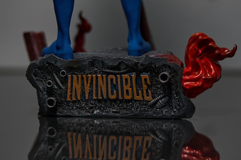 May include: A detailed statue base with the word "INVINCIBLE" in orange lettering. The base is dark gray with metallic accents and a red, flowing element on the side. Blue boots are visible above the base.