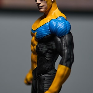 May include: A figurine of a superhero in a yellow and blue costume with black accents. The figure has a muscular physique, a yellow mask, and black hair. The costume features a star emblem on the chest.