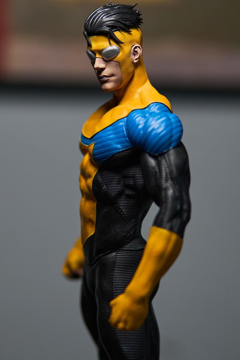 May include: A figurine of a superhero in a yellow and blue costume with black accents. The figure has a muscular physique, a yellow mask, and black hair. The costume features a star emblem on the chest.