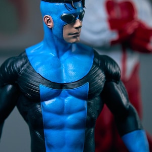 May include: A detailed action figure of a superhero in a blue and black suit with a mask. The figure has a muscular physique and is posed dynamically. Another figure is blurred in the background.
