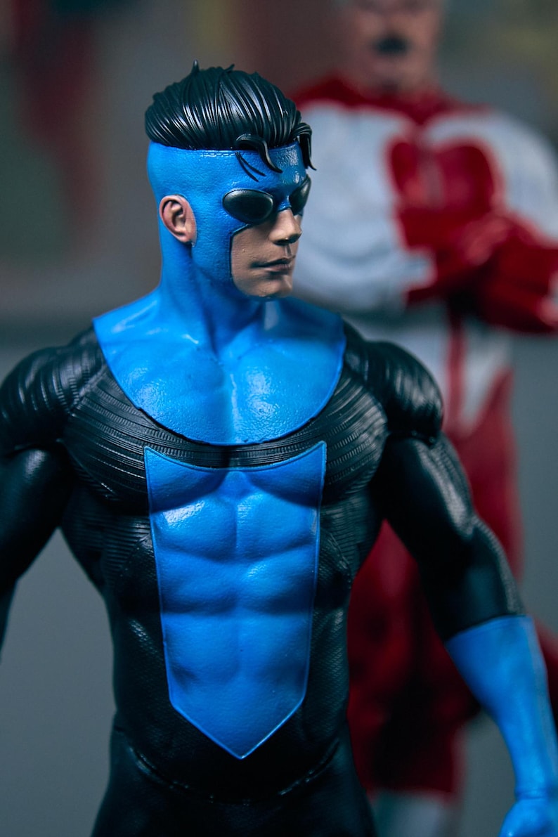 May include: A detailed action figure of a superhero in a blue and black suit with a mask. The figure has a muscular physique and is posed dynamically. Another figure is blurred in the background.