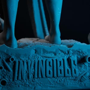 May include: A blue figurine base with the word "INVINCIBLE" embossed on it. The base has a textured surface with mechanical details and supports the legs of a figure. The background is dark.