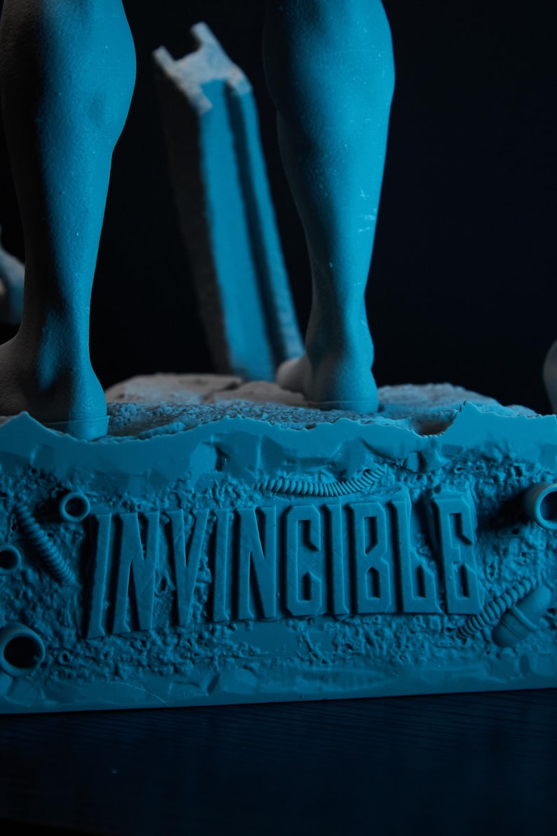 May include: A blue figurine base with the word "INVINCIBLE" embossed on it. The base has a textured surface with mechanical details and supports the legs of a figure. The background is dark.