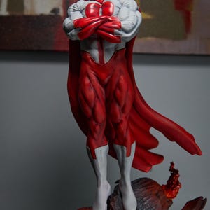 May include: A detailed figurine of a superhero in a dynamic pose. The figure is predominantly red and white, with a red cape and muscular physique. The character has a distinctive mustache and is standing on a rocky base.