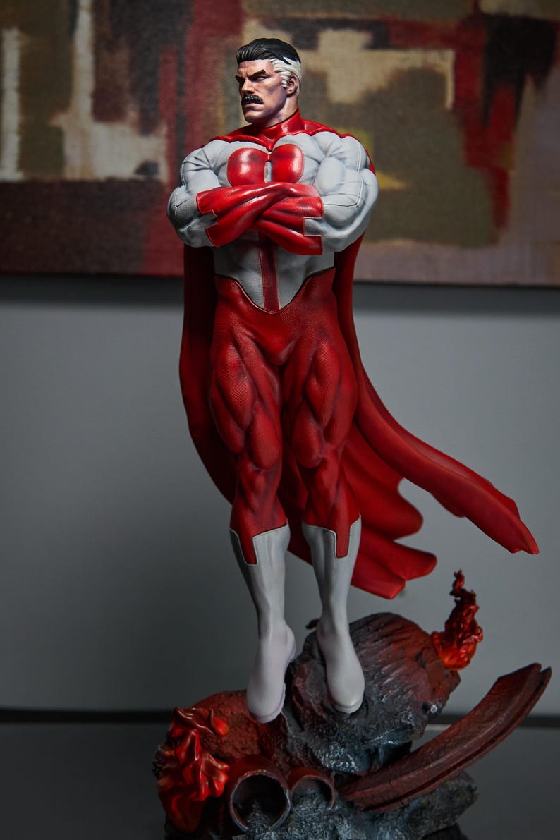 May include: A detailed figurine of a superhero in a dynamic pose. The figure is predominantly red and white, with a red cape and muscular physique. The character has a distinctive mustache and is standing on a rocky base.