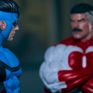 May include: Two action figures of superheroes. One figure is predominantly blue with black accents and a mask. The other figure is red and white with a black mustache. The figures are facing each other.