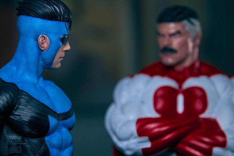 May include: Two action figures of superheroes. One figure is predominantly blue with black accents and a mask. The other figure is red and white with a black mustache. The figures are facing each other.