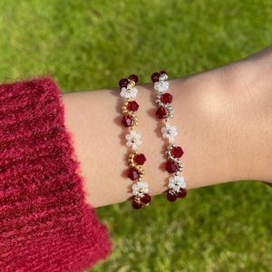 May include: Two beaded bracelets with red, white, and gold accents. One bracelet features gold beads and white flower-shaped beads, while the other has silver beads. Both have red crystal beads. The bracelets are worn on a wrist.