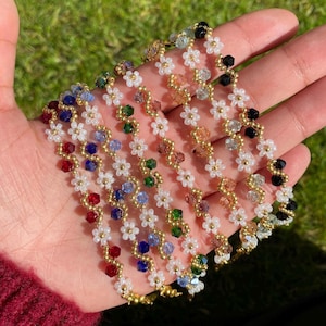 Op de afbeelding: Een set van zeven kralenkettingen met witte bloemenbedels en gekleurde kristallen kralen. De kettingen zijn waaiervormig op een hand gerangschikt.