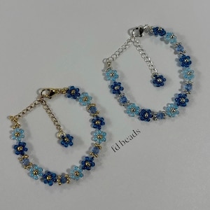 May include: Two beaded bracelets with a floral design. The bracelets feature light blue and dark blue flower beads with gold-coloured accents. Each bracelet has a chain extender and a clasp.