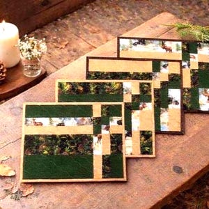 May include: A set of four rectangular fabric placemats featuring a patchwork design of dark green solid fabric, tan fabric, and a printed pattern depicting deer in a forest setting. The placemats are arranged in a staggered, overlapping stack.