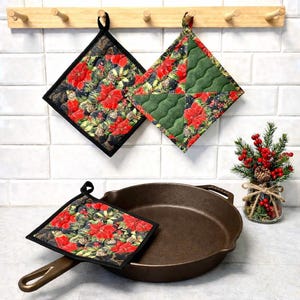 May include: A cast iron frying pan with a potholder featuring a red poinsettia and pinecone pattern. Two potholders hang on a wooden rack, and a small vase with berries and pinecones is on the counter.