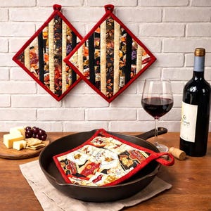 May include: Two quilted pot holders with a red border and a wine-themed fabric design hang on a white brick wall. A cast iron skillet holds a matching pot holder. A bottle of red wine, a wine glass, cheese, and grapes are also present.