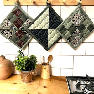 Op de afbeelding: Drie gewatteerde pannenlappen met ophanglussen. De pannenlappen zijn ruitvormig en hebben verschillende patronen, waaronder paisley en geometrische ontwerpen in groen, bruin en zwart. Ze hangen tegen een witte betegelde muur boven een houten aanrecht.