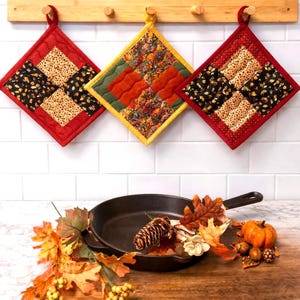 May include: Three quilted pot holders in red, yellow, and green hang from a wooden rack. Each pot holder features a patchwork design with autumn-themed fabrics. A cast iron skillet is decorated with fall leaves, a pinecone, and a small pumpkin.