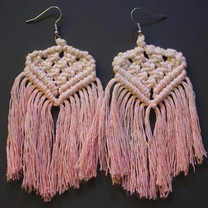 May include: A pair of pink and white macrame earrings with fringe. The earrings are made with a knotting technique and have a geometric design.