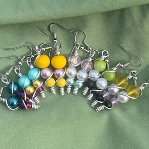 May include: A collection of handmade earrings with a variety of colorful beads. Each earring features a unique combination of colors, including yellow, blue, purple, and green, with silver wire detailing and fish hook closures.