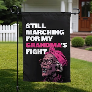 Può includere: Bandiera da giardino nera con la scritta "STILL MARCHING FOR MY GRANDMA'S FIGHT" in bianco e rosa. La bandiera presenta un'illustrazione di un'anziana signora con occhiali e un foulard rosa.