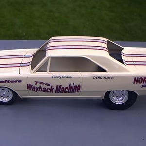 May include: A cream-colored model car with red and blue stripes on the roof. The car has the words "RaceCrafters," "The Wayback Machine," "Dyno Tuned," and "Norman Racing" printed on the sides. The car has black tires and silver rims.