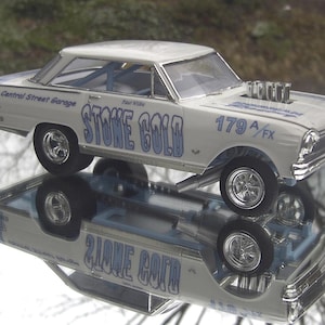 Può includere: Un modello di auto da corsa vintage, principalmente bianco con dettagli blu. L'auto presenta il testo "STONE COLD" e "179 A/FX" in blu. Sono visibili anche le parole "Central Street Garage" e "427 Dyno Tuned". L'auto si riflette su una superficie a specchio.