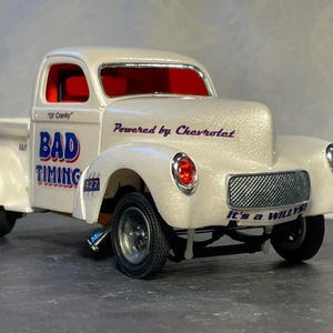 May include: A white vintage pickup truck model with the words "BAD TIMING" and "It's a WILLYS!" in blue. The truck has red taillights and a chrome grill. The text "Powered by Chevrolet" is also visible.