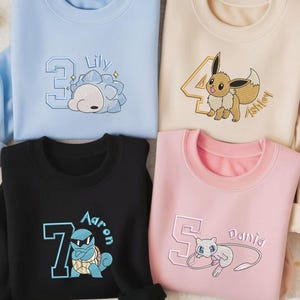 May include: Four sweatshirts in light blue, beige, black, and pink. Each features a number, a name, and a cartoon character embroidery. The light blue sweatshirt has a number 3 and the name "Lily". The beige sweatshirt has a number 4 and the name "Ashley". The black sweatshirt has a number 7 and the name "Aaron". The pink sweatshirt has a number 5 and the name "Dania".