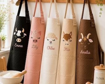 May include: Five aprons hanging on a wooden rack, each featuring an embroidered animal face and a name: Emma on black, Lily on pink, Chloe on cream, Mia on tan, and Amelia on brown. Each apron has a front pocket and adjustable button straps.