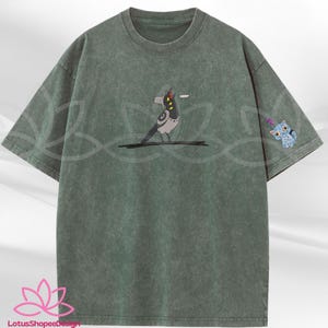 May include: Green t-shirt with a graphic of a bird with a speech bubble and a small blue cat on the sleeve. The shirt has a crew neck and short sleeves. The bird is gray, black, and white.