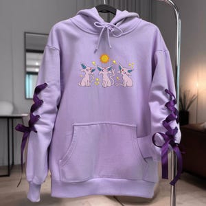 May include: A light purple hooded sweatshirt featuring a graphic of three Espeon characters and a yellow sun. The sleeves are accented with dark purple ribbon lacing that ties into bows at the cuffs. The hoodie includes a front kangaroo pocket.