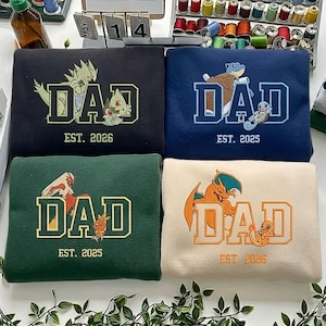 May include: Four sweatshirts with the word "DAD" in a collegiate font, each with a different Pokémon graphic and "EST. 2025" or "EST. 2026". The sweatshirts are black, navy blue, green, and cream-colored.