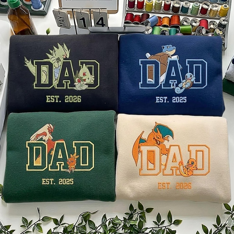 May include: Four embroidered sweatshirts in black, navy, dark green, and cream. Each features the word DAD in block letters with a Pok&eacute;mon character integrated into the design, accompanied by the text EST. 2025 or EST. 2026 below the word.