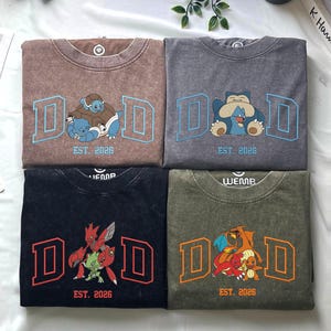 Op de afbeelding: Vier T-shirts met ronde hals in bruin, grijs, zwart en olijfgroen. Elk shirt heeft een grote letter D aan beide kanten van een Pokémon-afbeelding en de tekst EST. 2026. De afbeeldingen tonen Squirtle en Blastoise, Snorlax en Munchlax, Scizor en Scyther, en Charizard en Charmander.