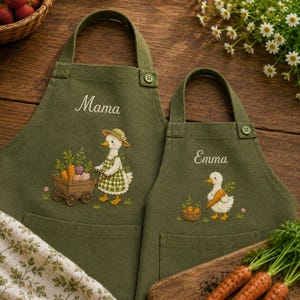Könnte beinhalten: Zwei olivgrüne Schürzen mit gestickten Entenmotiven. Die größere Schürze ist mit dem Wort Mama und einer Ente, die einen Karren mit Karotten zieht, bestickt. Die kleinere Schürze zeigt den Namen Emma und eine Ente, die eine Karotte hält.