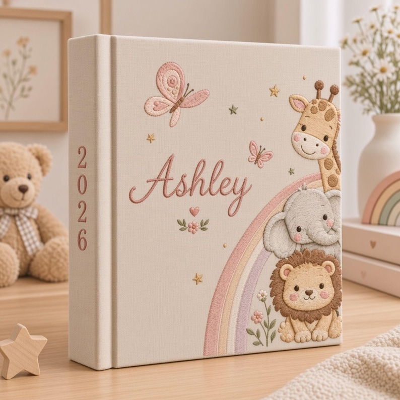 May include: A cream-colored fabric photo album featuring embroidered illustrations of a giraffe, elephant, and lion, along with a rainbow, butterflies, and stars. The name Ashley is embroidered in script, and the year 2026 is on the spine.