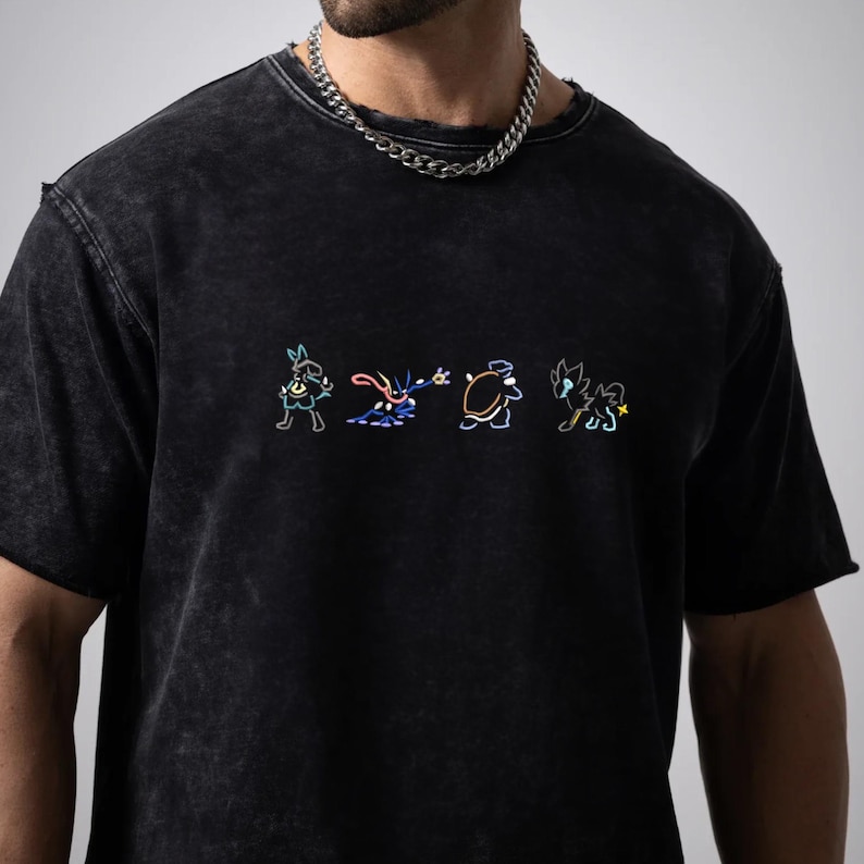 May include: Black, short-sleeved t-shirt with a worn appearance. The shirt displays a colourful embroidered design of four cartoon characters. The design is outlined in neon colours, creating a vibrant contrast against the dark fabric.
