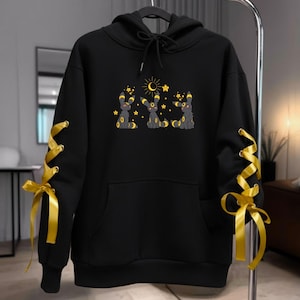 May include: Black hooded sweatshirt featuring a graphic of three cartoon characters with yellow accents, stars, and a crescent moon. The sleeves have yellow ribbon lacing details. The sweatshirt has a front pocket and a drawstring hood.