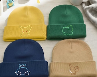 May include: Four colorful knit beanies, each with a different embroidered cartoon character. The beanies are yellow, green, navy blue, and tan. A coffee cup and small figurines are in the background.