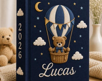 May include: A navy blue fabric-covered photo album featuring an embroidered hot air balloon with a teddy bear in the basket. The cover displays the name Lucas in white script, the year 2026 on the spine, and embroidered clouds, stars, and a moon.