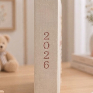 May include: A beige fabric-covered book spine featuring the year 2026 embroidered in a vertical orientation with rose-colored thread. The texture of the linen-like material is visible, and the numbers are stacked neatly in a simple sans-serif font.