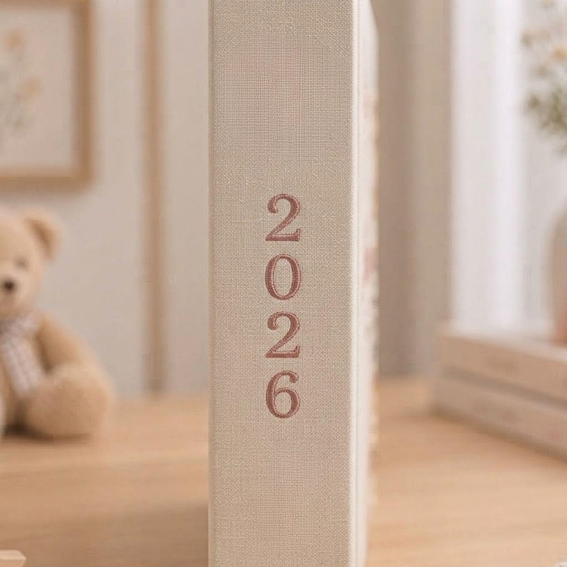 May include: A beige fabric-covered book spine featuring the year 2026 embroidered in a vertical orientation with rose-colored thread. The texture of the linen-like material is visible, and the numbers are stacked neatly in a simple sans-serif font.