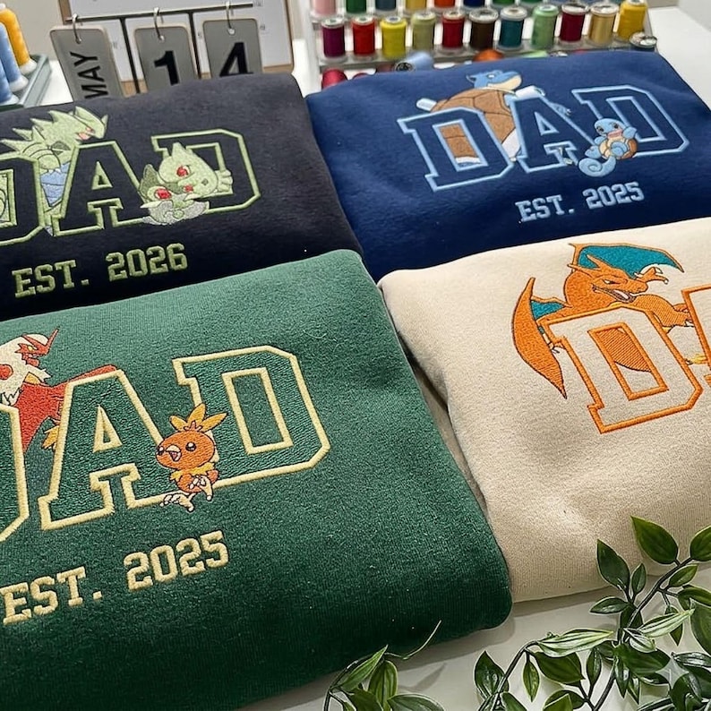 May include: Four embroidered sweatshirts in black, navy blue, dark green, and cream. Each features the word DAD in large block letters with various Pok&eacute;mon characters, including Blastoise, Squirtle, and Charizard, plus EST. 2025 or 2026 text.