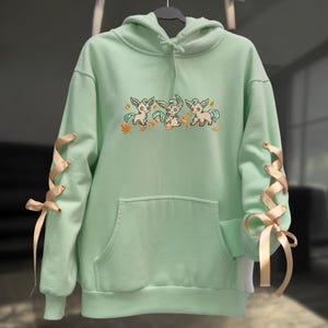 May include: A mint green hooded sweatshirt featuring a central embroidered graphic of three small, light-colored creatures surrounded by orange autumn leaves. The long sleeves are accented with beige ribbon lace-up details tied into bows.