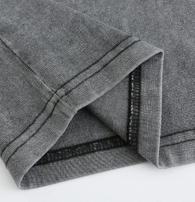 May include: Close-up of a gray fabric garment, possibly a t-shirt or shirt, with visible seams and stitching. The fabric has a textured appearance, and the seams are reinforced with black thread. The garment is folded, revealing the inside.