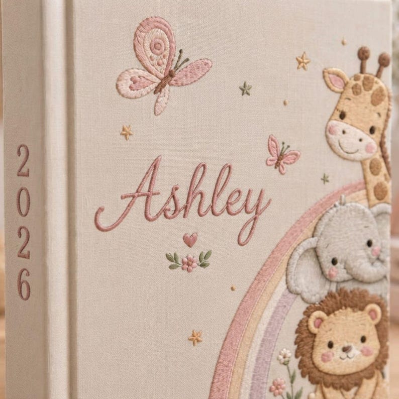 May include: A beige fabric-covered book featuring embroidered designs of a butterfly, a giraffe, an elephant, and a lion. A rainbow arc is stitched below the animals. The name Ashley is embroidered in pink script, and the year 2026 is on the spine.