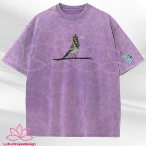 May include: A purple t-shirt with a unique design. The front features a cartoon bird-like character with a red hat. The left sleeve has a small blue owl graphic. The shirt has a textured, washed appearance.